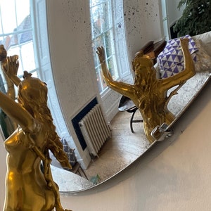 May include: A gold-colored statue of a woman with her arms raised, reflected in a round, slightly distorted mirror. The mirror is mounted on a white wall.