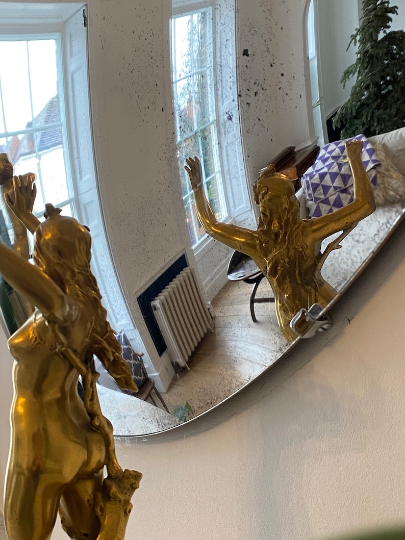 May include: A gold-colored statue of a woman with her arms raised, reflected in a round, slightly distorted mirror. The mirror is mounted on a white wall.