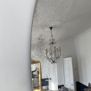 May include: A large, oval-shaped antique mirror with a distressed silver finish. The mirror reflects a room with a white ceiling, a chandelier, and a couch with pillows.