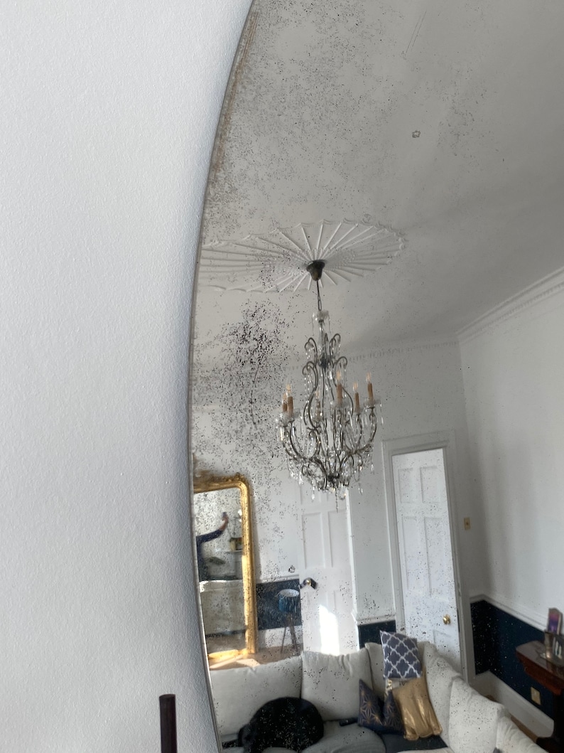 May include: A large, oval-shaped antique mirror with a distressed silver finish. The mirror reflects a room with a white ceiling, a chandelier, and a couch with pillows.