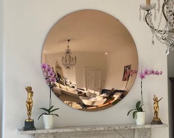 May include: A large, round, copper-toned mirror hangs above a white marble fireplace. Two small potted orchid plants with pink flowers are on either side of the fireplace. Two gold statues are also on the mantel.