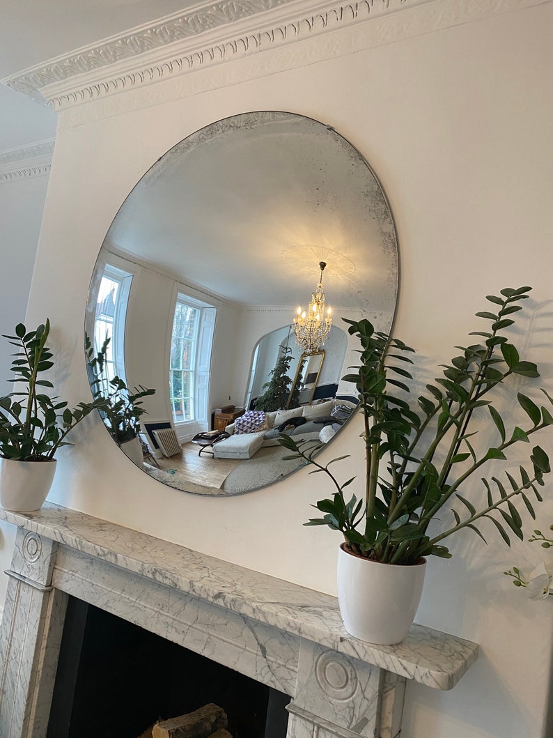 May include: A large, round, antique mirror with a silver frame hangs above a white marble fireplace mantel. Two potted plants with green leaves are on either side of the mirror.