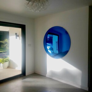 Puede incluir: Un gran espejo de pared convexo, circular y de tono azul, montado en una pared blanca. La superficie del espejo refleja la habitaci&oacute;n circundante, incluyendo una puerta y luz natural, creando una vista distorsionada de gran angular del espacio.