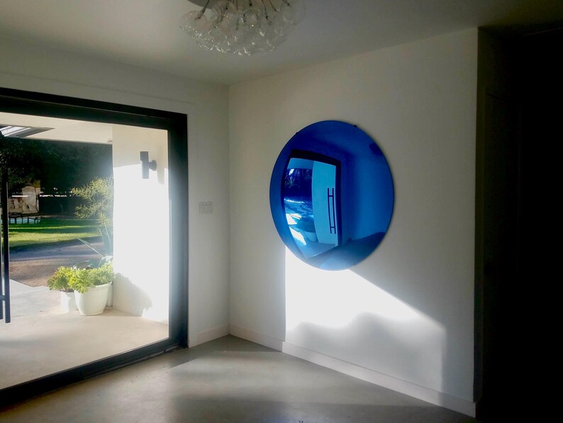 Puede incluir: Un gran espejo de pared convexo, circular y de tono azul, montado en una pared blanca. La superficie del espejo refleja la habitaci&oacute;n circundante, incluyendo una puerta y luz natural, creando una vista distorsionada de gran angular del espacio.