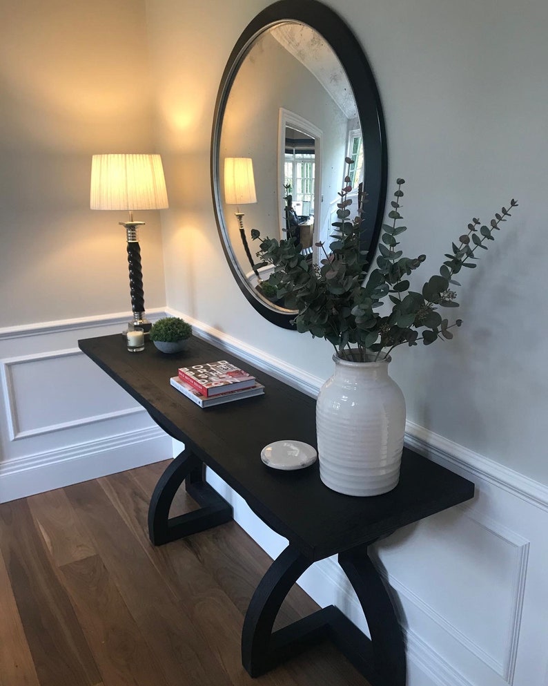 May include: A dark wood console table with a curved base, holding a white vase with greenery, a round mirror, a lamp, and decorative items. The table is made of wood and has a dark finish. The mirror has a black frame.