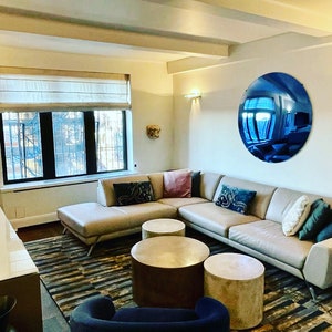 Puede incluir: Una sala de estar con un gran sof&aacute; seccional beige con varios cojines, tres mesas de centro cil&iacute;ndricas con acabados en oro met&aacute;lico, una alfombra oscura estampada y un espejo de pared circular azul que refleja el interior de la estancia.