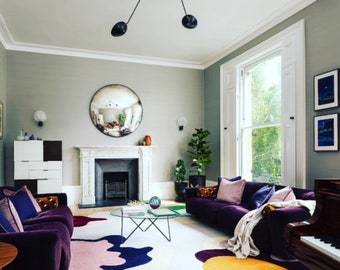 Pode incluir: Uma sala de estar com um sofá de veludo roxo, um tapete branco com formas abstratas coloridas, uma lareira com uma grelha preta e um grande espelho redondo acima da lareira.