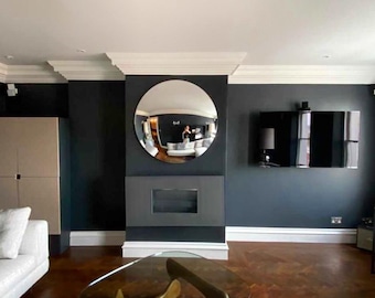 May include: A living room with dark walls, a white ceiling, and a large, round, silver mirror. The mirror is hanging above a fireplace. There is a glass coffee table in the center of the room.
