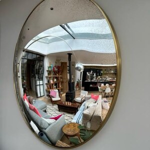 Pode incluir: Um espelho redondo com moldura dourada refletindo uma sala de estar com um sof&aacute;, cadeiras e uma lareira. O espelho est&aacute; montado em uma parede cinza.
