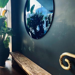 Puede incluir: Un gran espejo de pared circular con marco oscuro montado en una pared gris oscuro sobre un banco de madera largo y desgastado. A la derecha, un pasamanos de metal dorado pulido con un extremo curvo en espiral fijado a la pared.