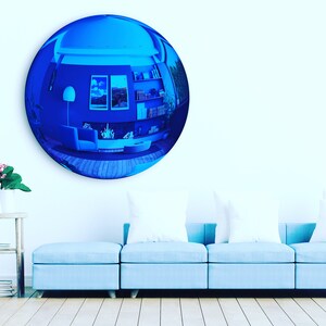 Puede incluir: Un gran espejo de pared convexo, circular y con tinte azul que refleja el interior de una habitaci&oacute;n con un sof&aacute;, estanter&iacute;as y cuadros. El espejo tiene una superficie lisa y reflectante que distorsiona la vista en una perspectiva gran angular.