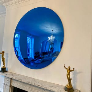 Puede incluir: Un gran espejo circular con una superficie de cristal tintado en azul intenso. El espejo refleja el interior de una habitaci&oacute;n, incluyendo una l&aacute;mpara de ara&ntilde;a, ventanas y muebles. Est&aacute; montado en una pared sobre una repisa de m&aacute;rmol con dos estatuas doradas.