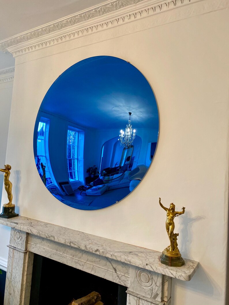 Puede incluir: Un gran espejo circular con una superficie de cristal tintado en azul intenso. El espejo refleja el interior de una habitaci&oacute;n, incluyendo una l&aacute;mpara de ara&ntilde;a, ventanas y muebles. Est&aacute; montado en una pared sobre una repisa de m&aacute;rmol con dos estatuas doradas.