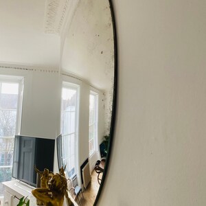 May include: A large, oval-shaped antique mirror with a silver finish. The mirror is mounted on a white wall and reflects a room with windows and furniture.
