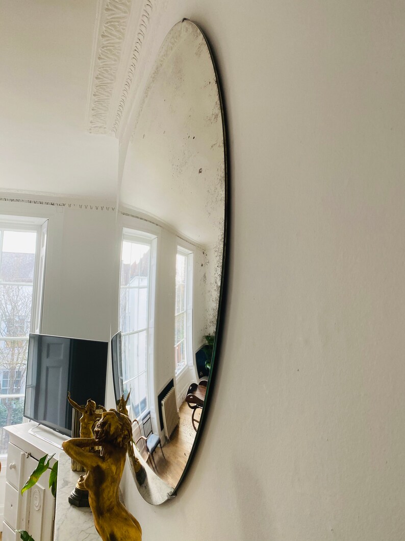 May include: A large, oval-shaped antique mirror with a silver finish. The mirror is mounted on a white wall and reflects a room with windows and furniture.