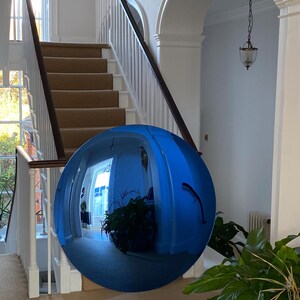 Puede incluir: Un espejo convexo circular grande con un tinte azul y una superficie altamente reflectante. El espejo muestra un reflejo distorsionado de gran angular de un espacio interior, que incluye una escalera, paredes blancas y plantas verdes.