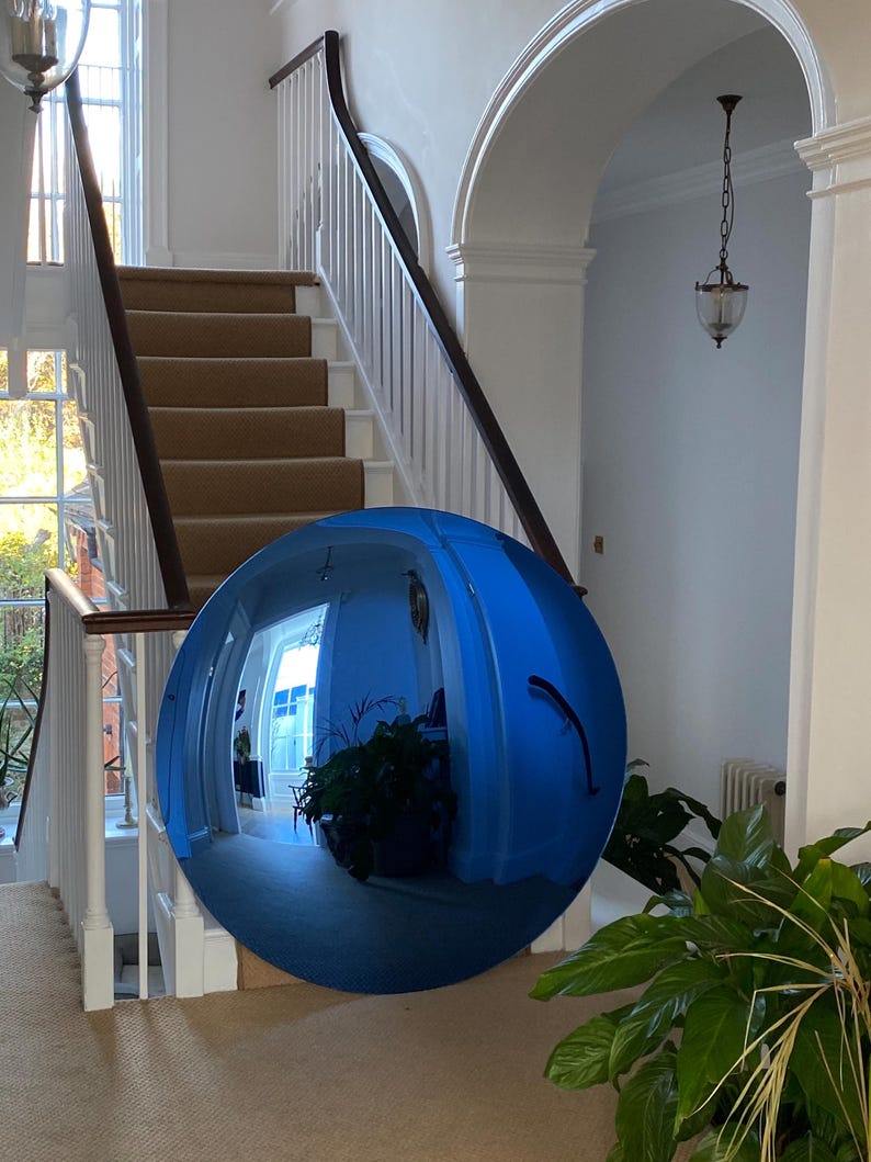 Puede incluir: Un espejo convexo circular grande con un tinte azul y una superficie altamente reflectante. El espejo muestra un reflejo distorsionado de gran angular de un espacio interior, que incluye una escalera, paredes blancas y plantas verdes.