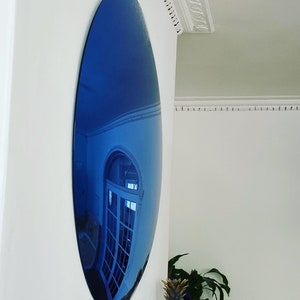 Puede incluir: Un espejo convexo circular grande con tinte azul montado en una pared blanca. La superficie del espejo es altamente reflectante y muestra un reflejo azul profundo y distorsionado de un espacio interior con una puerta blanca acristalada.