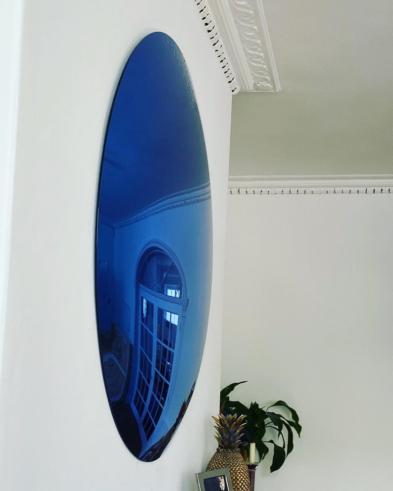 Puede incluir: Un espejo convexo circular grande con tinte azul montado en una pared blanca. La superficie del espejo es altamente reflectante y muestra un reflejo azul profundo y distorsionado de un espacio interior con una puerta blanca acristalada.