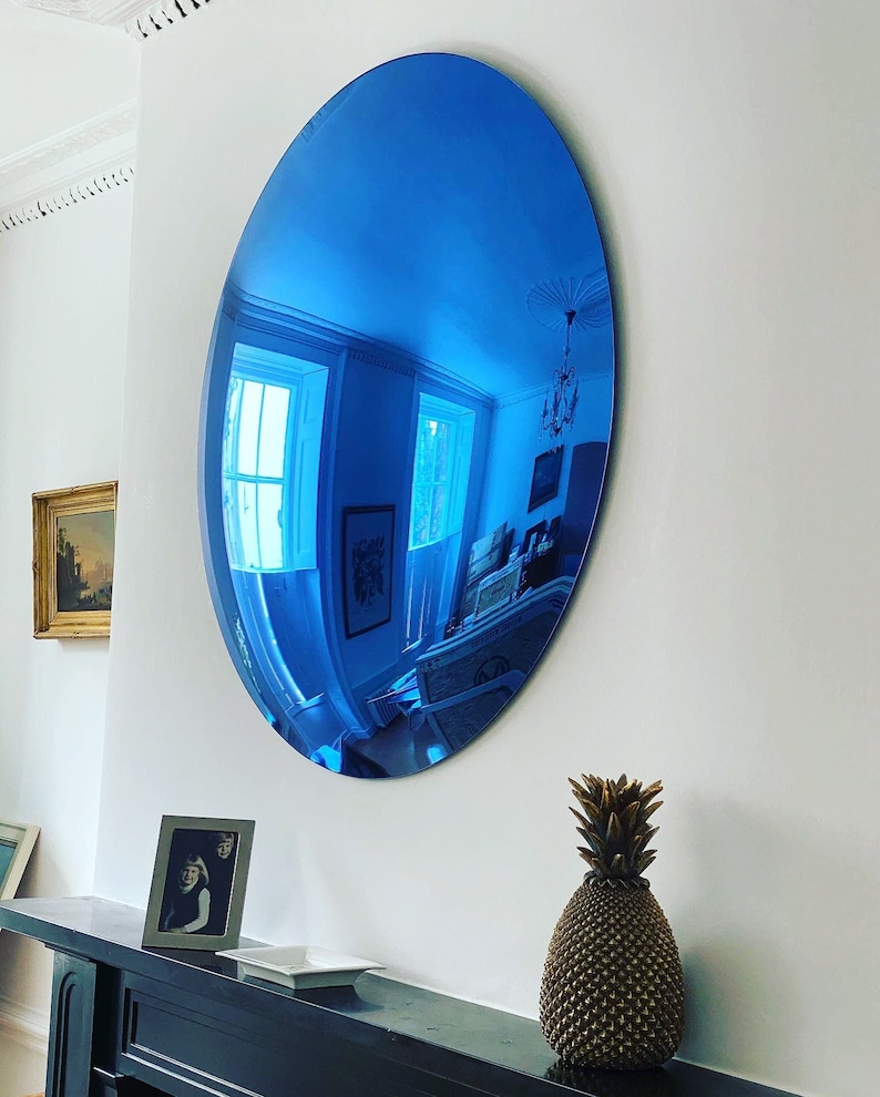 Puede incluir: Un gran espejo de pared de forma ovalada con una superficie reflectante de color azul vibrante. El espejo est&aacute; montado en una pared blanca lisa, creando un reflejo azul profundo y distorsionado del interior, incluyendo ventanas y l&aacute;mparas.