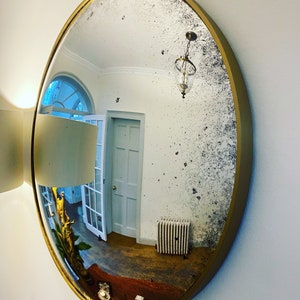 Pu&ograve; includere: Uno specchio grande, rotondo, con cornice dorata e superficie invecchiata dall'aspetto antico. Lo specchio riflette una stanza con soffitto bianco, pareti bianche e una porta bianca.