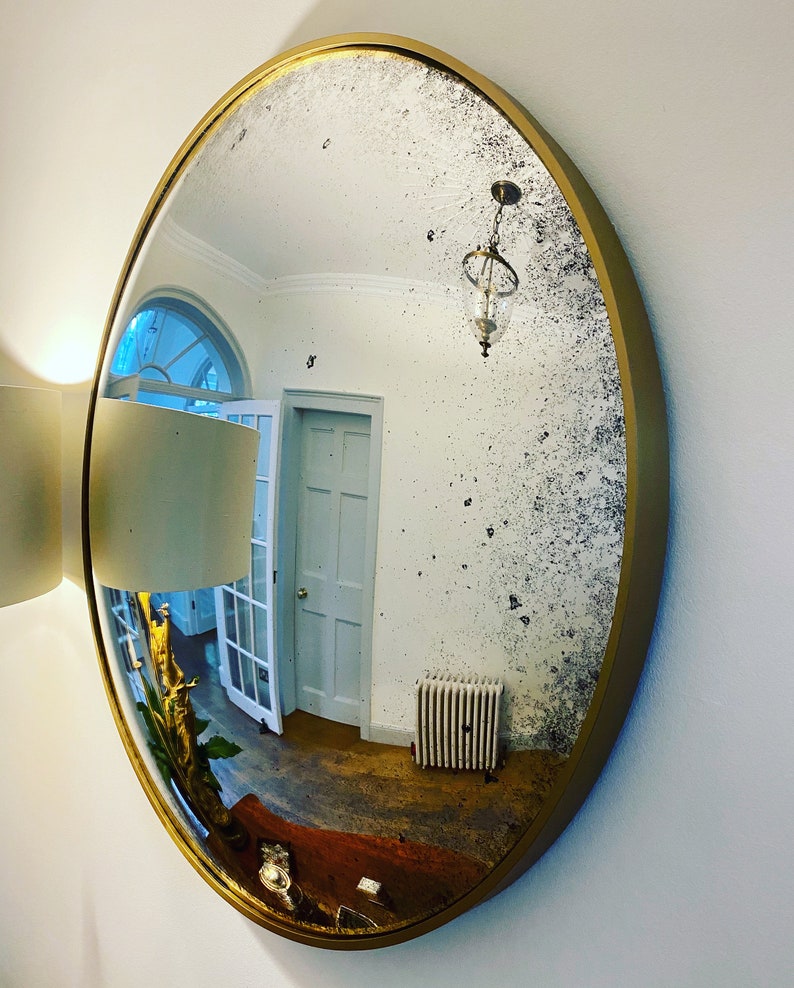 Pu&ograve; includere: Uno specchio grande, rotondo, con cornice dorata e superficie invecchiata dall'aspetto antico. Lo specchio riflette una stanza con soffitto bianco, pareti bianche e una porta bianca.