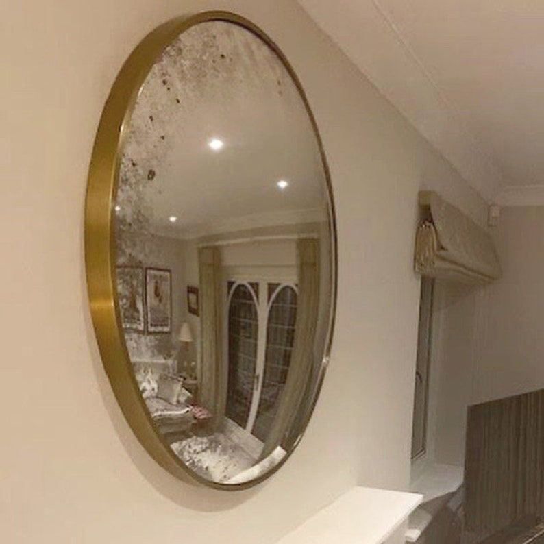 Pode incluir: Um espelho grande, redondo, com moldura dourada e um acabamento ligeiramente desgastado. O espelho reflete uma sala com uma porta e cortinas.