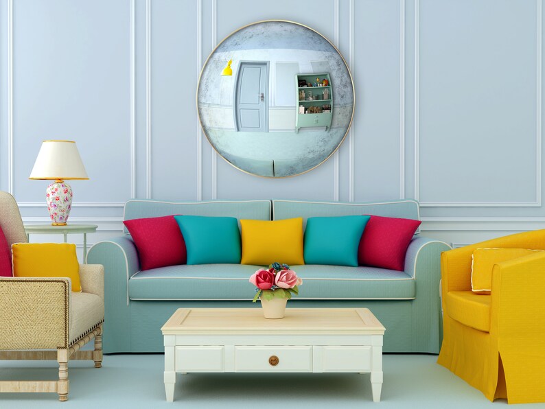 Pode incluir: Um interior de sala de estar com um sof&aacute; azul claro, duas poltronas, uma mesa de centro e um grande espelho redondo na parede. O sof&aacute; tem almofadas coloridas em tons de vermelho, amarelo, azul e turquesa. As poltronas s&atilde;o amarelas e bege. A mesa de centro &eacute; branca com uma gaveta. H&aacute; flores em um vaso sobre a mesa de centro.