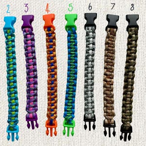 May include: Nine colorful paracord bracelets with plastic buckles are displayed. The bracelets feature various color combinations, including pink, blue, purple, orange, green, gray, brown, and olive green. Each bracelet is approximately 0.75 inches wide.