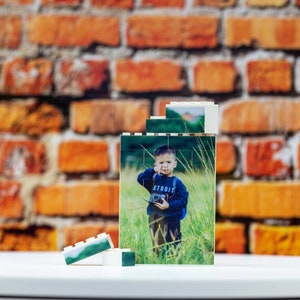 Op de afbeelding: Een wit Lego-model van een jonge jongen die een camera vasthoudt in een grasveld. Het model is gemaakt van verschillende kleinere stenen en heeft een geprinte afbeelding van de jongen.