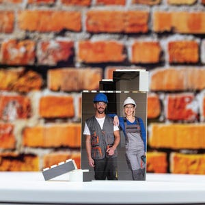 May include: Two people wearing hard hats and work clothes stand in front of a brick wall. The image is printed on a white rectangular block.