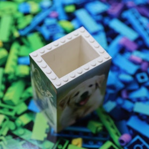 May include: White Lego brick pencil holder with a picture of a golden retriever dog on the front. The holder is surrounded by colorful Lego bricks.