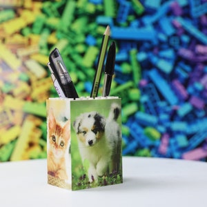 May include: A white ceramic pencil holder with a colorful background. The holder features a cat and a dog on the front. There are three pens and a pencil in the holder.