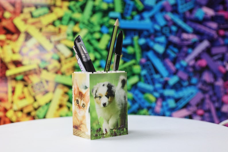 May include: A white ceramic pencil holder with a colorful background. The holder features a cat and a dog on the front. There are three pens and a pencil in the holder.