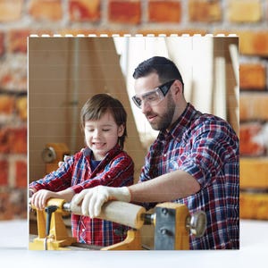 May include: A father and son are working together in a woodshop. The father is wearing safety glasses and a plaid shirt. The son is wearing a red and black plaid shirt. They are using a wood lathe to shape a piece of wood.