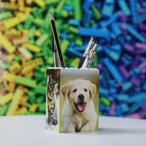 May include: White plastic pencil holder with a cat and a puppy on the sides. The holder is filled with pens and pencils. The background is a blurred image of colorful plastic bricks.
