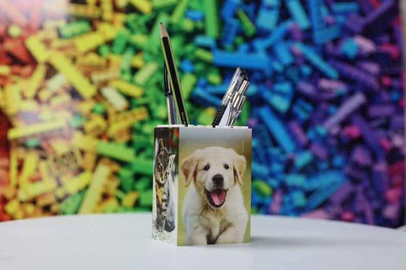 May include: White plastic pencil holder with a cat and a puppy on the sides. The holder is filled with pens and pencils. The background is a blurred image of colorful plastic bricks.