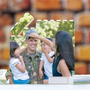 May include: A family portrait featuring a man in military uniform, a woman, and two children. The man is smiling and holding a young girl in his arms. The woman is smiling and placing her hand on the man's head. The children are smiling and looking at the camera. The background is a blurred image of green trees and foliage.