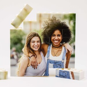 Op de afbeelding: Twee vriendinnen lachen naar de camera, hun gezichten worden omlijst door een witte fotopuzzel. De puzzel is gemaakt van afzonderlijke vierkante stukjes die in elkaar passen om een ​​groter beeld te creëren. De foto toont de vriendinnen in een parksetting.