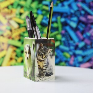 May include: A white ceramic pencil holder with a photo of a puppy and a kitten on the front. The holder is filled with pens and pencils.