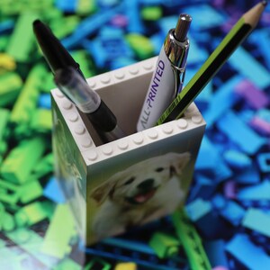 May include: A white Lego pencil holder with a picture of a puppy on the front. The holder has the words "All-Printed" on the side. There are two pencils and a pen inside the holder.