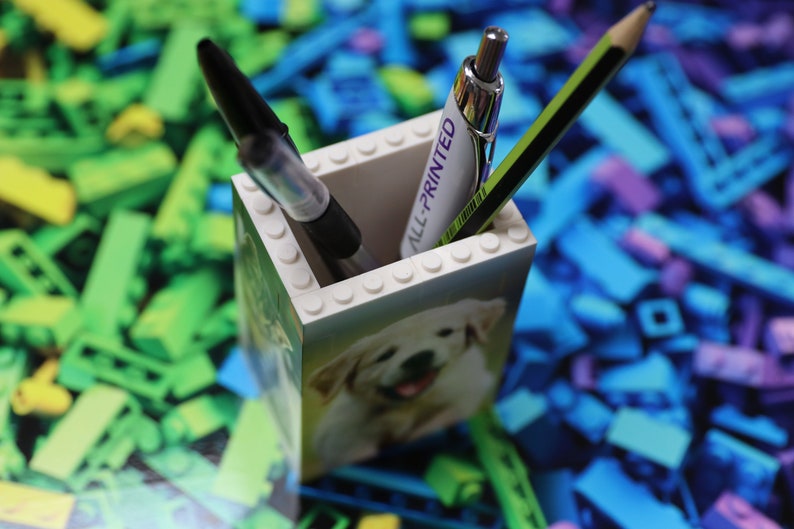 May include: A white Lego pencil holder with a picture of a puppy on the front. The holder has the words "All-Printed" on the side. There are two pencils and a pen inside the holder.