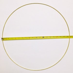 May include: A gold-coloured metal ring, approximately 50 cm in diameter, is shown against a white background. A yellow measuring tape is placed across the ring, indicating its size. The ring is thin and appears to be made of wire.