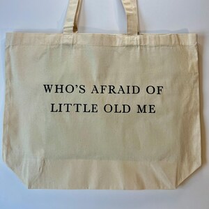 May include: A beige canvas tote bag with long handles. The bag features the text "WHO'S AFRAID OF LITTLE OLD ME" printed in black. The bag is rectangular and has a simple, minimalist design.