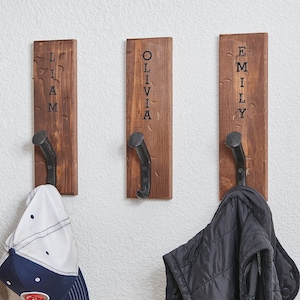 May include: Four wooden wall hooks with metal hooks, each with a name engraved on the front. The names are Mia, Liam, Olivia, and Emily. Two hats and a jacket are hanging on the hooks.