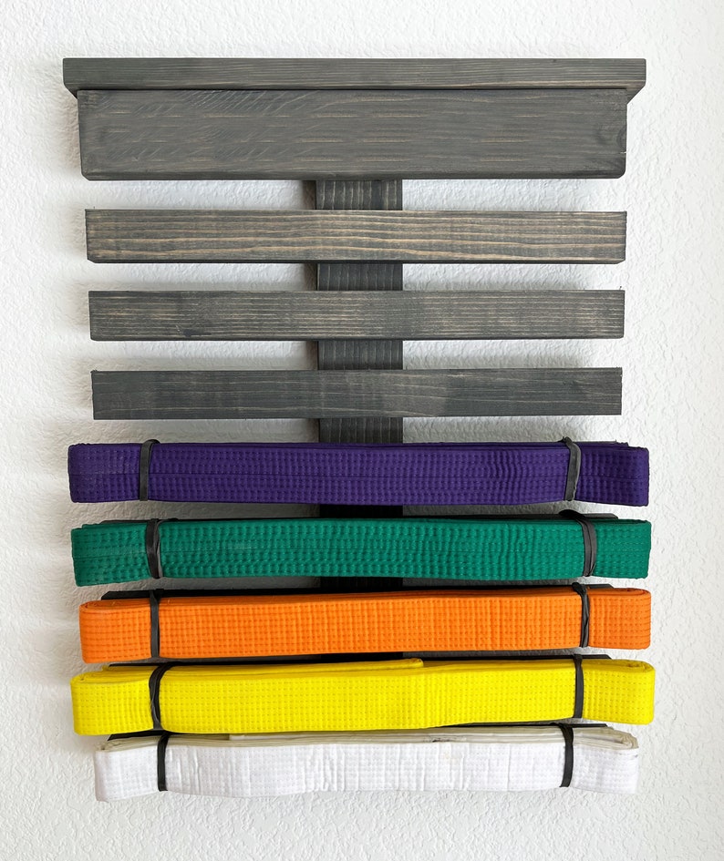 May include: A wooden martial arts belt display rack with a dark gray finish. The rack holds several colored belts: white, yellow, orange, green, and purple. Each belt is secured with a black band. The rack is mounted on a white wall.