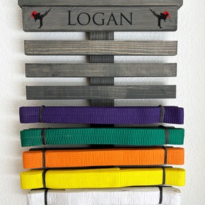 May include: A wooden martial arts belt display with the name "LOGAN" in black lettering. The display has multiple horizontal slats holding colored belts: purple, green, orange, yellow, and white. Karate figures are on each side of the name.