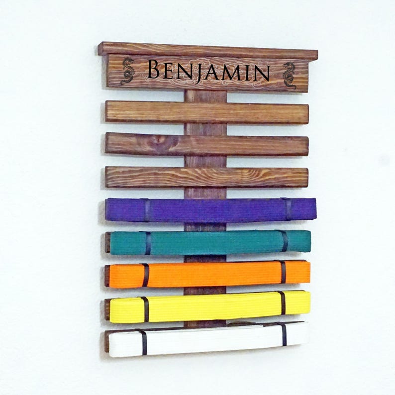 May include: A wooden wall-mounted belt rack with the name "BENJAMIN" engraved on the top. The rack has seven slots for belts, and there are six belts displayed in various colours, including purple, green, orange, yellow, and white.