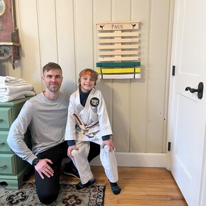 May include: A wooden belt display rack with the name "PAUL" above the slots. The rack holds green, yellow, and white belts. A person in a white karate uniform and a brown belt stands in front of the rack with another person.
