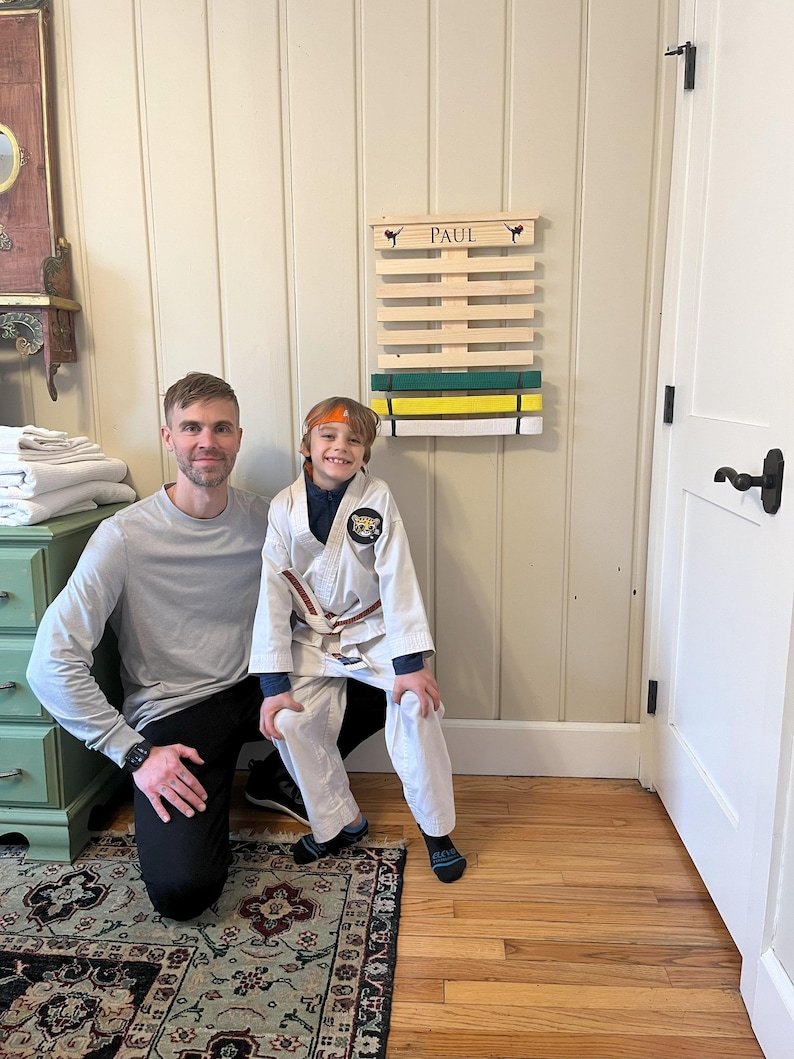 May include: A wooden belt display rack with the name "PAUL" above the slots. The rack holds green, yellow, and white belts. A person in a white karate uniform and a brown belt stands in front of the rack with another person.